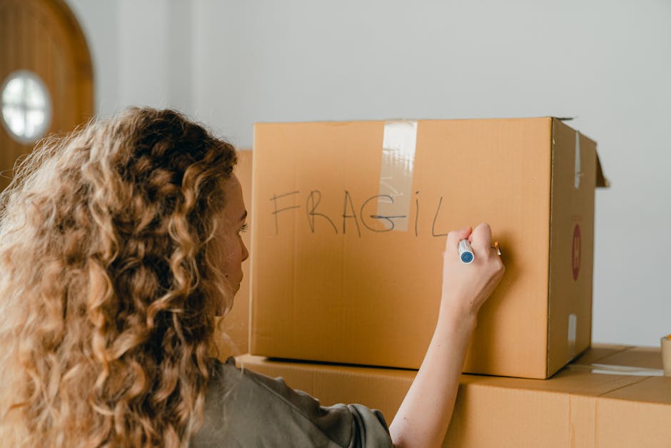 A woman with long, curly hair is inside a home, writing the word 'FRAGILE' in large letters on a cardboard moving box with a blue marker. The box is positioned on top of several other packed cardboard boxes arranged near a doorway. The setting appears to be a room with neutral-colored walls, and natural light is illuminating the scene. This image depicts an ongoing home relocation or packing process, which is part of the furniture transport and packing preparations handled by Finchley Man and Van, a local removals company specializing in house removals and moving services in Finchley.