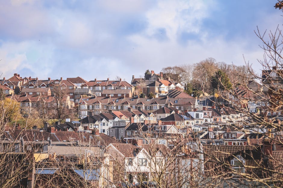 A panoramic view of a residential area featuring densely packed houses on a hillside. The scene includes rooftops made of red tiles, gray asphalt, and some flat roofs with chimneys. In the foreground, bare tree branches partially obscure the lower houses, indicating a season like late autumn or winter. The sky is partly cloudy with patches of blue sky and soft, diffused sunlight illuminating the scene. The area appears to be a typical suburban neighbourhood with a mix of detached and semi-detached homes, some with visible window frames, small front gardens, and driveways. The houses are situated on different levels of the slope, showing varied elevation. This environment suggests typical home relocation or moving context, with possible evidence of furniture or packing materials not visible in this specific image. The setting underscores professional house removals, such as those provided by Finchley Man and Van, in a well-established residential community.