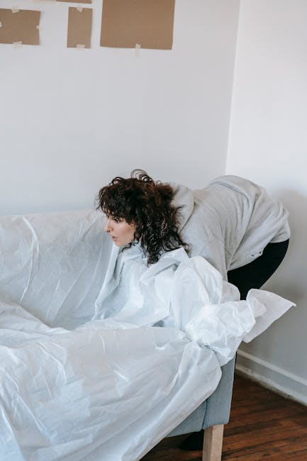 A woman with curly dark hair is seen bending over and lifting a large, white padded moving blanket that is draped over a grey fabric sofa inside a home. The scene is set in a well-lit room with white walls, one of which has three pieces of brown packing paper taped to it. She is wearing a light grey hoodie and dark trousers. In the background, the wall is plain and unadorned, and the floor appears to be made of dark wood. The sofa is partially covered with packing materials, possibly in preparation for a home relocation or furniture transport. This image depicts the packing or furniture moving process typical of professional removals services provided by Finchley Man and Van, who specialise in house removals near Stephens House in East Finchley, aligning with their services in packing, loading, and furniture transport.