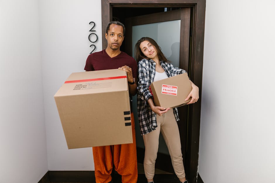 Within the interior of a residential property, several cardboard boxes and wrapped furniture pieces are visible, indicating an ongoing home relocation process. The boxes are stacked near a doorway or opening, some sealed with packing tape, while a large piece of furniture, possibly a wardrobe or cabinet, is covered with protective fabric or bubble wrap. On the floor, there are moving blankets and plastic wrapping materials used for furniture protection. In the background, a moving trolley and straps are seen, suggesting the transport of heavy items. The scene is illuminated by natural daylight, highlighting the organized packing and loading activities typical of professional removals. The environment appears clean and well-organized, consistent with the packing and furniture transport stages managed by Finchley Man and Van during their house removal service near East Finchley, close to Stephens House.