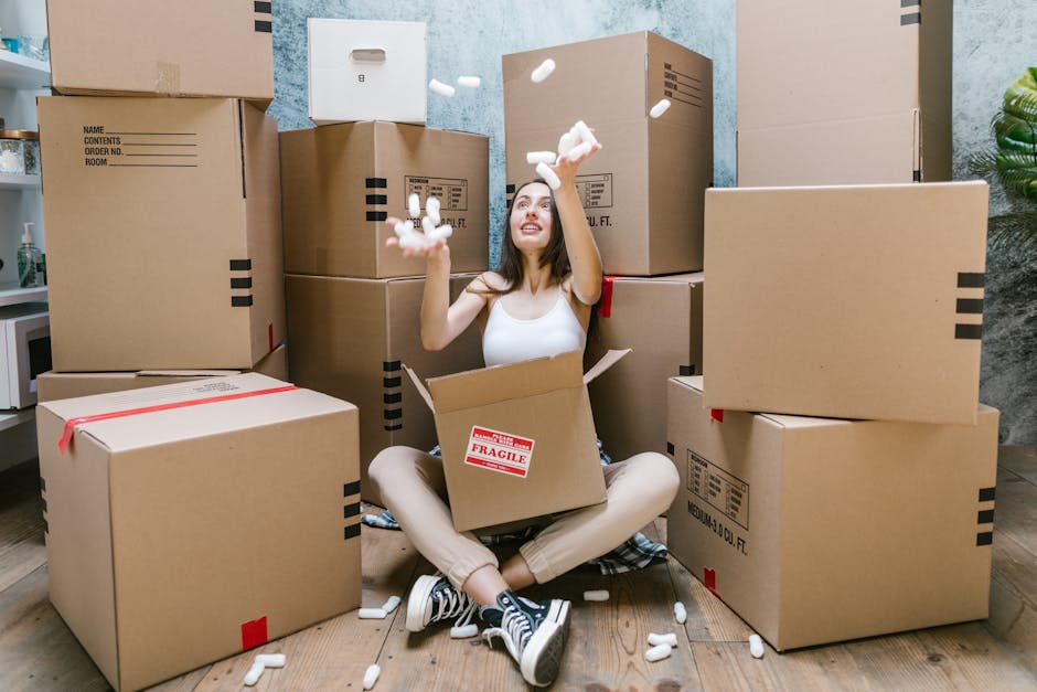 A young woman sitting cross-legged on a wooden floor surrounded by numerous large cardboard boxes during a home relocation process. She is inside a room with light-colored textured walls, and several of the boxes are stacked behind her and to her sides. Some boxes are sealed with red or black tape, while others are open, showing crumpled packing materials and foam packing peanuts falling from her hands. The woman is smiling and engaging with the packing materials, with a box labeled 'FRAGILE' positioned in her lap. In the background, a small white kitchen appliance and a green houseplant are visible on a counter, indicating the interior of a residential property. The scene reflects the packing and loadings phase of furniture transport or house removals, with natural indoor lighting illuminating the space and highlighting the logistics of home moving services as provided by Finchley Man and Van.