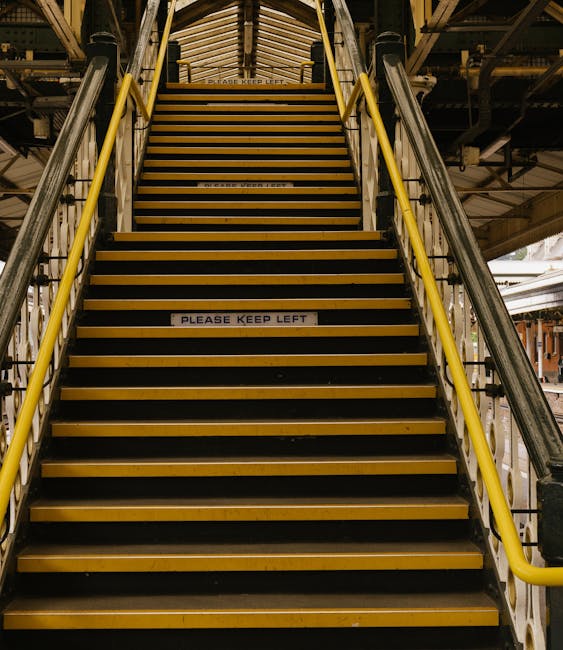 A set of industrial stairs inside a building, with yellow handrails on both sides and black and yellow striped anti-slip treads. The stairs lead upwards towards a platform with a warning sign visible
