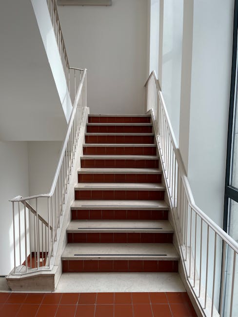 A set of industrial stairs inside a building, with yellow handrails on both sides and black and yellow striped anti-slip treads. The stairs lead upwards towards a platform with a warning sign visible