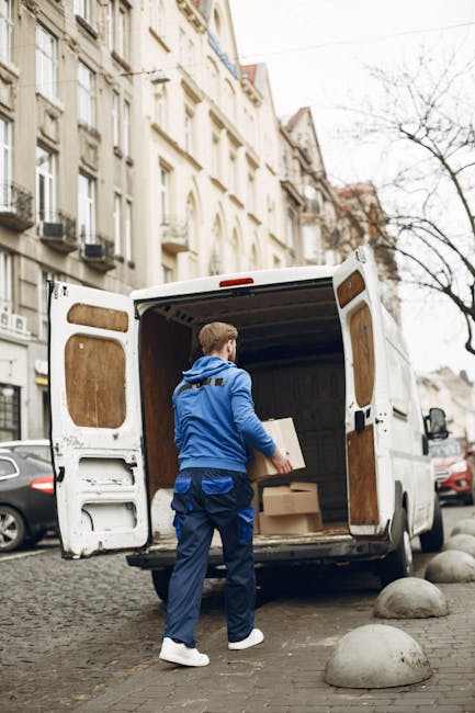 A person wearing a blue jacket and dark blue trousers is standing at the rear of a white box van, which is parked on a cobblestone street in an urban residential area. The van has open rear doors revealing an interior lined with wooden panels, and the person is carrying two cardboard boxes, one in each hand, preparing for a home relocation. The loading area is adjacent to a building with a classic façade featuring large windows and balconies, and there are several parked cars along the street. The scene includes a row of large concrete bollards on the pavement to prevent vehicle overrun, and leafless trees are visible in the background, indicating a colder season. This image exemplifies the packing and loading process during furniture transport for house removals, with the professional services of Finchley Man and Van supporting efficient moving logistics.