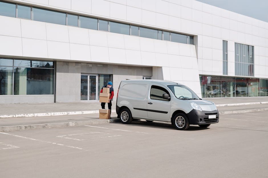 A person in a red jacket and blue beanie is seen standing outside a modern commercial building, unloading several cardboard boxes into a small, grey delivery van parked on a paved area. The boxes are stacked on the ground next to the individual, who appears to be in the process of loading or preparing for home relocation or furniture transport as part of a professional removals service by Finchley Man and Van. The van is positioned close to the building's entrance, which features large glass windows and a glass door, with the vehicle's side door open for access. The surroundings include a clean, well-marked parking area with designated spaces and minimal traffic, indicating an organized loading zone for moving purposes. The environment is well-lit, with natural daylight illuminating the scene, supporting a smooth loading process typical of local house removals and packing activities.