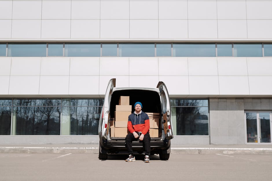A person wearing a blue jacket and dark blue trousers is standing at the rear of a white box van, which is parked on a cobblestone street in an urban residential area. The van has open rear doors revealing an interior lined with wooden panels, and the person is carrying two cardboard boxes, one in each hand, preparing for a home relocation. The loading area is adjacent to a building with a classic façade featuring large windows and balconies, and there are several parked cars along the street. The scene includes a row of large concrete bollards on the pavement to prevent vehicle overrun, and leafless trees are visible in the background, indicating a colder season. This image exemplifies the packing and loading process during furniture transport for house removals, with the professional services of Finchley Man and Van supporting efficient moving logistics.